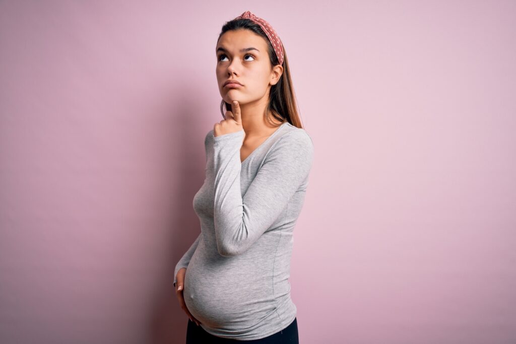 Young pregnant woman thinking, standing in front of pink background
